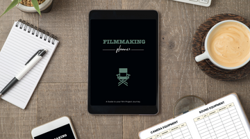 A digital filmmaking planner on a tablet, with a coffee mug, notepad, pen, plant and the ultimate filmmaking planner