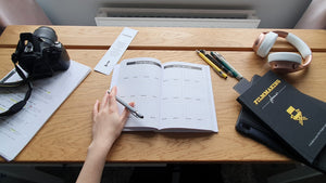 Filmmaking planner open on storyboard page with a camera and book mark. A hand hovers over the book with a pen. 